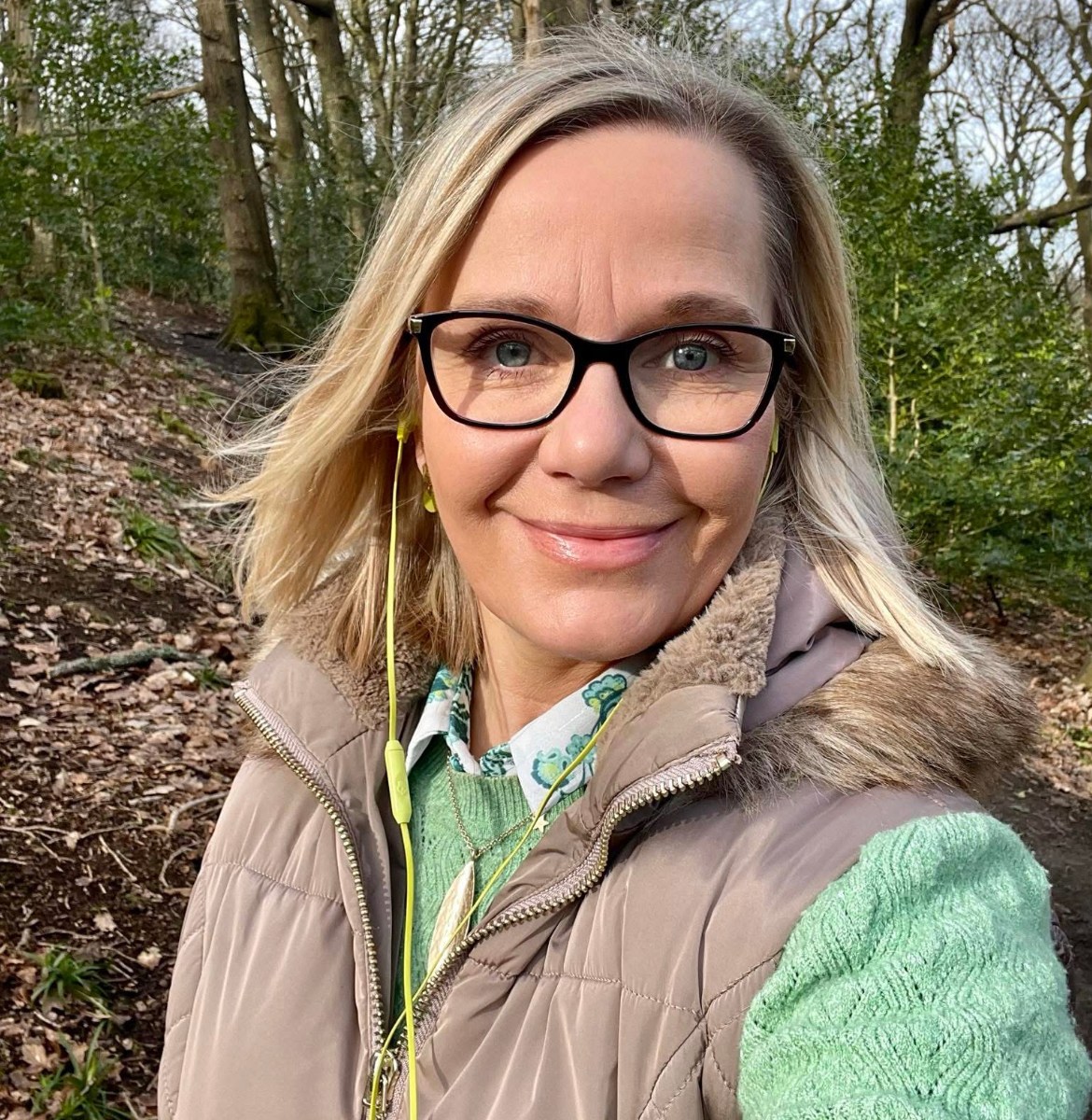 blonde woman wearing glasses and a green jumper, walking in the woods and smiling