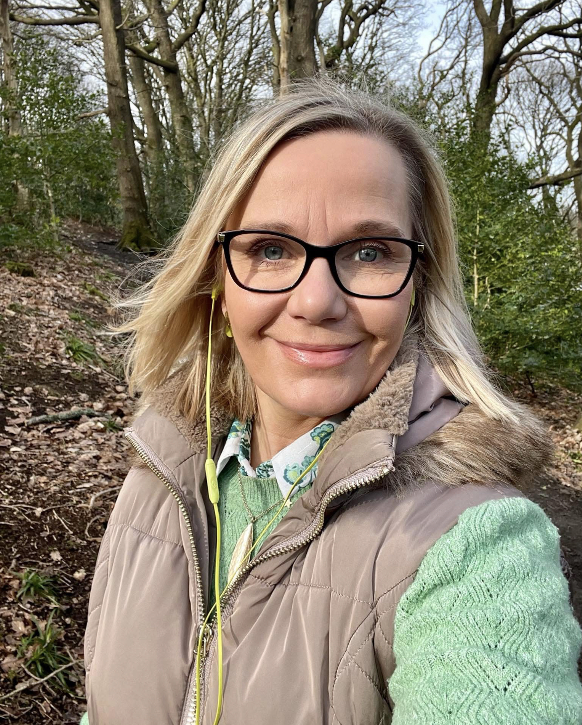 blonde woman wearing glasses and a green jumper, walking in the woods and smiling