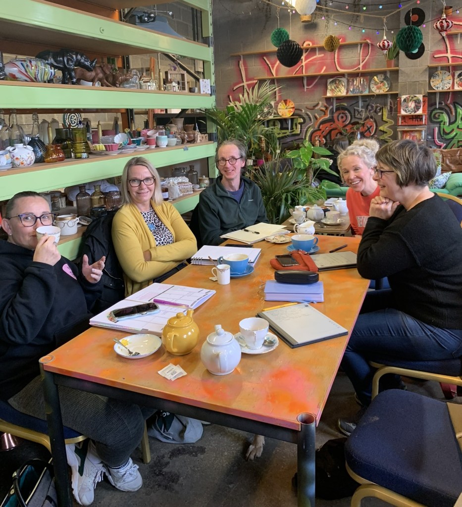 A group of 5 people sat around a table in a creative cafe smiling and drinking coffee